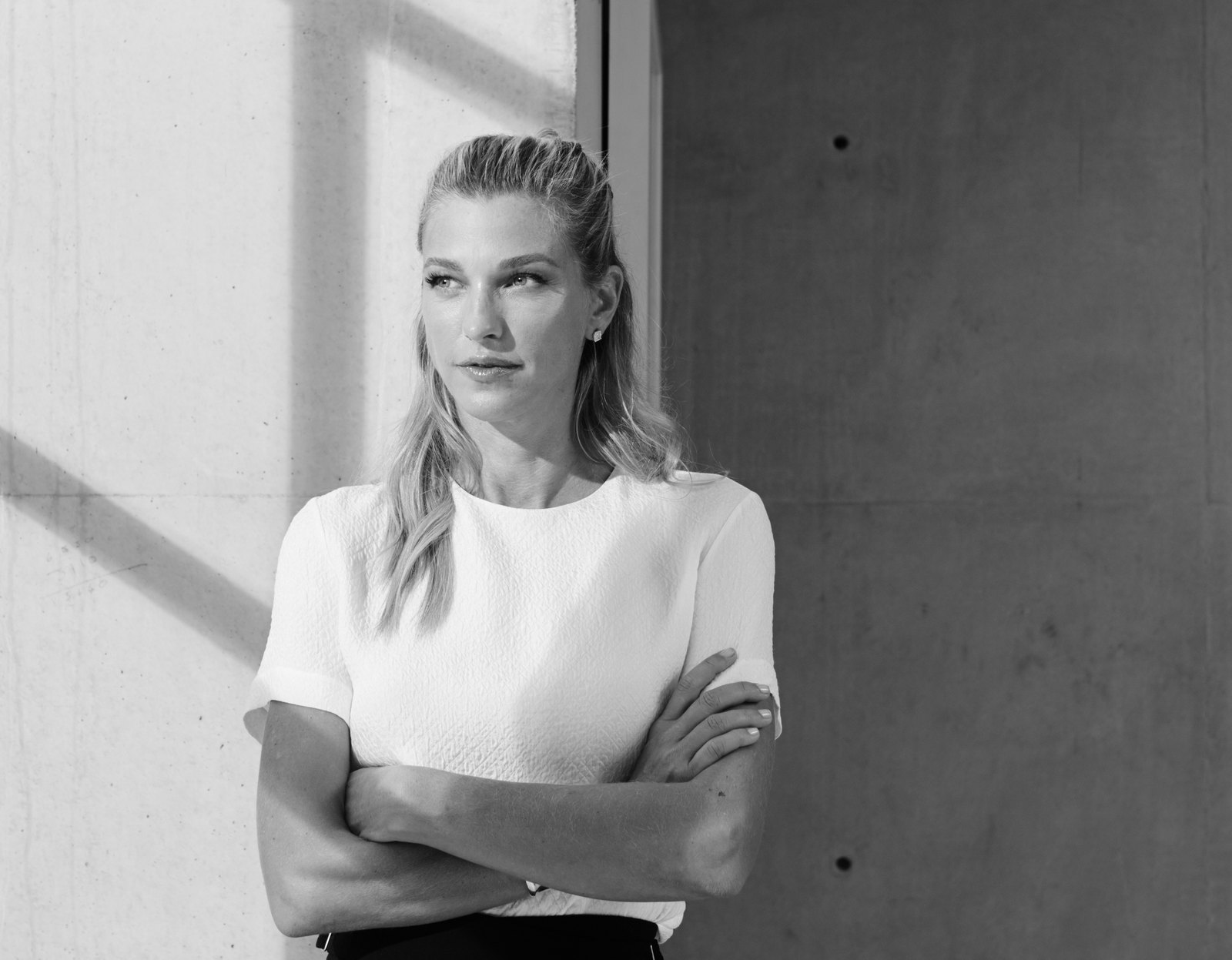 Nicole Junkermann in maglia bianca a braccia conserte accanto a una parete di cemento, in una fotografia in bianco e nero